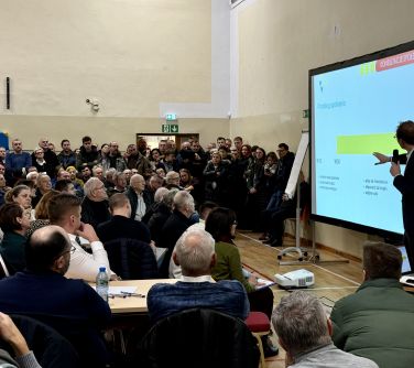 Oświetlona hala. Wiele osób słucha mężczyzne, który przemawia przy dużym ekranie, pokazując na wykres. Część siedzi, w tle...