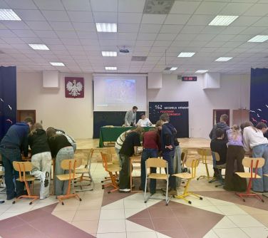 Jasna hala sportowa. Uczniowie pracują w grupach przy trzech stolikach. Na przodzie na podwyższeniu przy stole przykrytym...