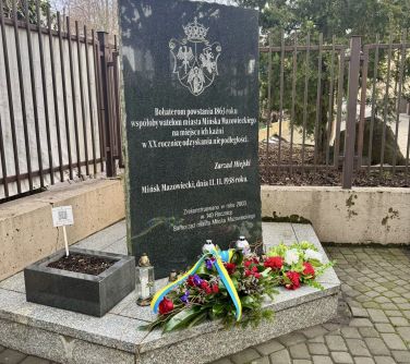 Dwór, dzień. Na betonowym wzniesieniu ciemna płyta jako pomnik bohaterów powstania styczniowego. Przed pomnikiem kwiaty