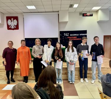 Jasna aula. Osiem uśmiechniętych osób stoi przed podwyższoną sceną. W tle wisi biały ekran