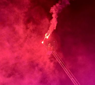 Wysunięty wysięgnik wozu strażackiego. W koszu strażak z dwiema odpalonymi racami. Niebo pokryte czerwonym dymem