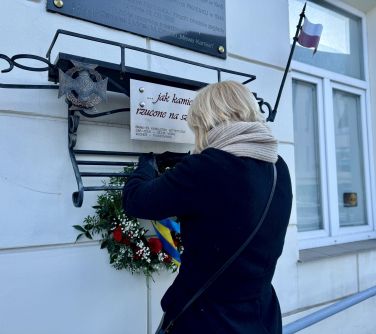 Dzień, na dworze. Przed białą ściana budynku kobieta w czarnym płaszczu wiesza okrągłą wiązankę na metalowym zaczepie