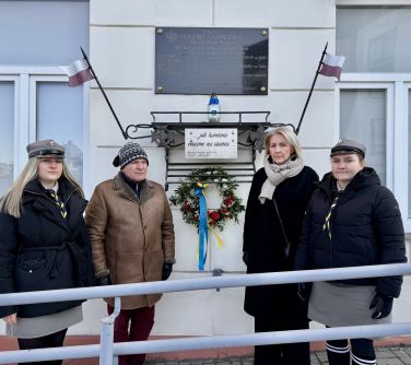 Na dworze, dzień. Przed białą ścianą budynku cztery osoby pozują do zdjęcia. Na ścianie metalowa tablica i zawieszka