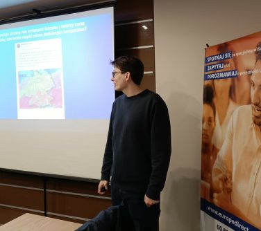 Sala konferencyjna, dzień. Po prawej baner z mężczyzną. Mężczyzna w ciemnym ubraniu patrzy na wyświetlana treść na ekranie