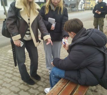 Na dworze, dzień. Na przystanku dwie nastolatki wręczają kolorową wlepke chłopcu siedzącego na ławce