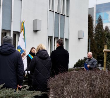 Pochmurny dzień. Przed jasnym budynkiem osoby, sztandar. Po prawej do mikrofonu przemawia mężczyzna