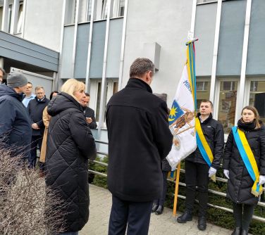 Pochmurny dzień. Przed jasnym budynkiem osoby oraz trzy osoby z pocztem sztandarowym