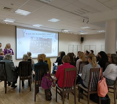 Hala konferencyjna. Na krzesłach siedza osoby. na przodzie ekran z treścią. Po lewej stoja dwie kobiety