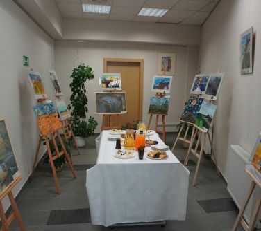 Oświetlone pomieszczenie. Po środku stół z białym obrusem. na stole poczęstunek. Naokoło na sztalugach prace plastyczne