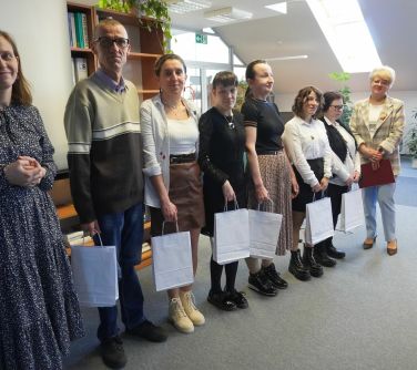 Oświetlone pomieszczenie. Po lewej osiem osób. Sześć z nich w środku trzyma białe torby papierowe