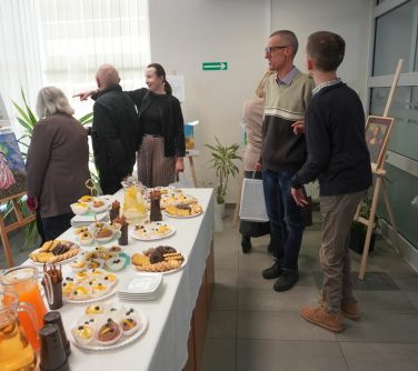 Oświetlone pomieszczenie. Grupa osób ogląda prace plastyczne położone na drewnianych sztalugach. Na środku stół z białym...