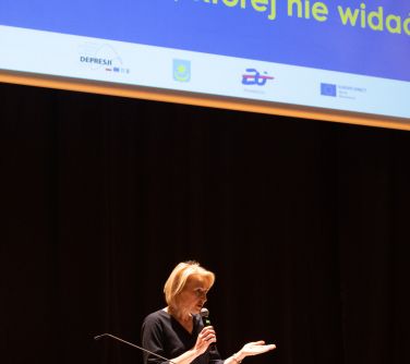 Przed mównicą kobieta przemawia do mikrofonu. W tle na dole czarna kotara, na górze wyświetlany tekst na ekranie