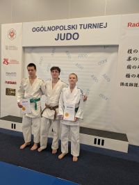 Oświetlone pomieszczenie. Trzech zawodników w białych kimonach stoi przed podium. W tle biała ścianka z informacjami i loga.