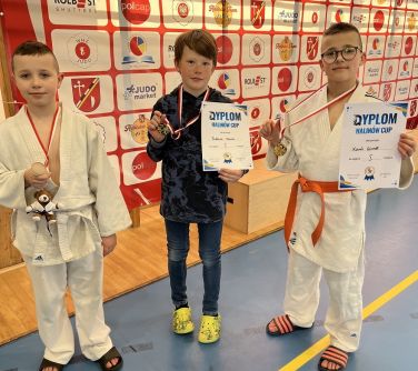 Oświetlona hala sportowa. Trzech chłopców z medalami. Dwóch trzyma również dyplomy. Stoją na niebieskiej podłodze. W tle...