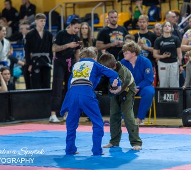 Oświetlona hala sportowa. Na niebiesko-różowej piankowej macie walczy dwóch chłopców. W tle kibice