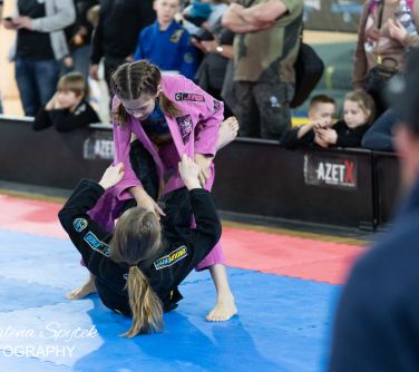 Hala sportowa. Na niebieskich piankowych matach walczą dwie nastolatki. Jedna w czarnym stroju w pozycji połleżącej. Nad...