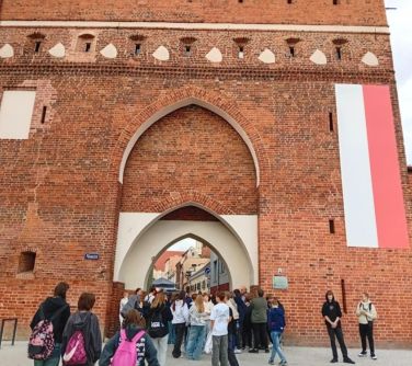 Dzień. przed wysokim murem z bramą grupa nastolatków. Po prawej stronie na ścianie długa flaga biało-czerwona