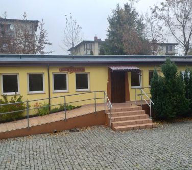 Na zewnątrz, dzień. zdjęcie niskiego, żółtego budynku. W tle drzewa i bloki