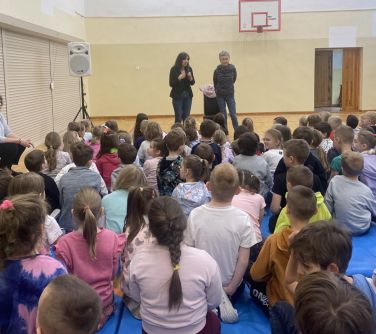 Oświetlona hala sportowa. Na podłodze siedzi grupa dzieci. Na przodzie dwie kobiety
