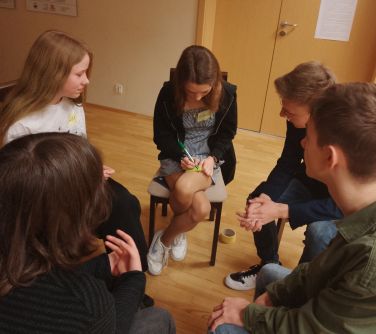 Sala. W kole na krzesłach młodzież. Jedna dziewczyna na kolanie pisze na żółtej kartce