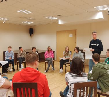 Oświetlona aula. W kole na krzesłach mlodzież. Po prawej stoi chłopak w czarnej bluzie z dużą kartką w dloni