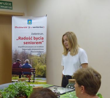 Oświetlone pomieszczenie.Przy stole z zielonym obrusem seniorzy. Na stole roślinki. Na przodzie kobieta w białej bluzce