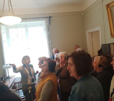 Oświetlone wnętrze. Seniorzy słuchaja kobiety. W tle okno, dzień