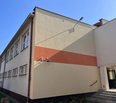 Na zewnątrz, dzień. Widok na jasna ścianę budynku z paśkiem pomarańczowym na środku