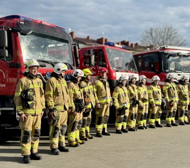 Na placu, dzien. Grupa strażaków w strojach roboczych. Stoja przed wozami strażackimi