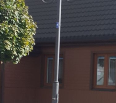 Na dworze, dzień. Latarnia z dwoma kloszami po bokach. W tle drewniany, niski budynek