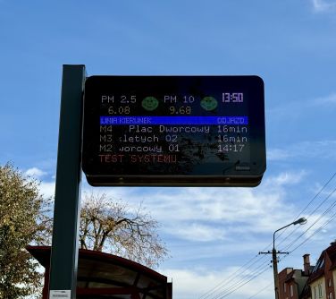 Na dworze, sloneczny dzień. Widok na tablicę elektryczną z wyświetlanym rozkładem jazdy autobusów. W tle błękitne niebo.
