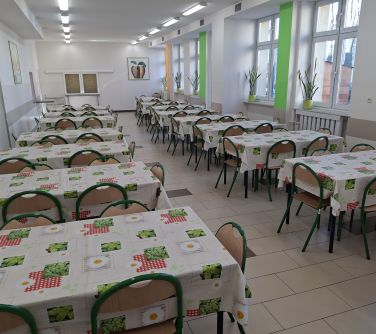 Oświetlona stołówka szkolna. W dwóch rzędach ustawione stoły. Przykryte bezowymi ceratami w kwiaty i liście w kwadratach...