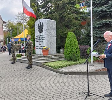 Na dworze, dzień. Po prawej mężczyzna w garniturze przemawia do stojącego mikrofonu. Po lewej dwóch żołnierzy przed pomnikiem