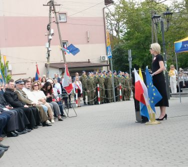 Na dworze, dzień. Kobieta w czarnej sukience przemawia przy mównicy. Obok flagi. na przodzie słuchacze
