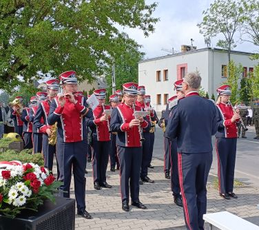 Na dworze, dzień. Na chodniku gra orkiestra w odświętnych garniturach. Po lewej na kolumnie wiązanka z biało-czerwonych...