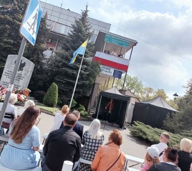 Na dworze, dzień. Widok na budynek. Na barierce flaga Polski. Za flagą trzech przebranych mężczyzn. Na dole na krzesłach...