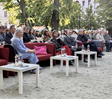 Na dworze, dzień. Na czerwonych kanapach, za nimi na krzesłach siedzą osoby. Przed kanapami trzy białe kwadratowe soliki