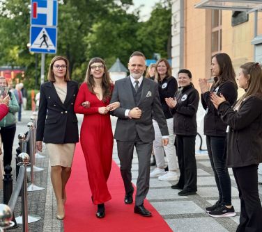 Na dworze, dzień. Dwie kobiety i mężczyzna po prawej maszerują pod ręcę. Idą po czerwonym dywanie