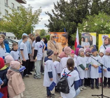 Na zewnątrz. Grupa przedszkolaków w białych koszulkach śpiewa do mikrofonu. Za nimi stoją ludzie.