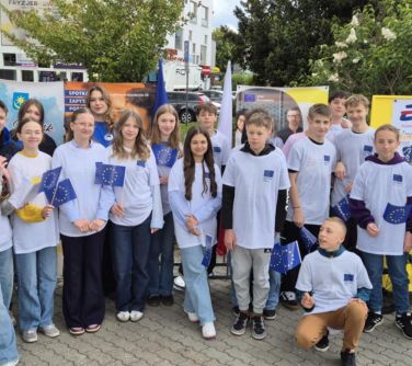 Na zewnątrz. Grupa młodzieży w białych koszulkach pozuje do zdjęcia. Towarzyszy im kobieta w płaszczu.