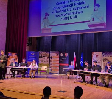 Widok na scenę w sali koncertowej. Młodzież siedzi przy dwóch stołach, chłopak stoi przy mównicy. W tle rollupy.
