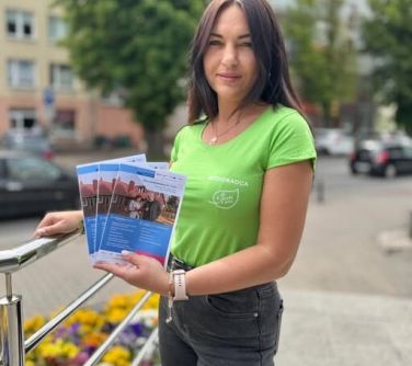 Na dworze, dzień. Zdjęcie kobiety w zielonej podkoszulce. Trzyma w dłoni trzy ulotki