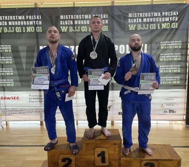 Oświetlona hala sportowa. Na podium trzech mężczyzn z dyplomami i medalami na szyi.