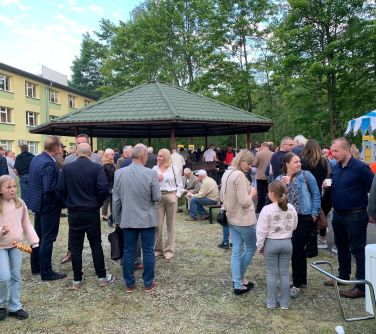 Na dworze. Pochmurny dzień. Spotkanie grupy osób. Część osób trzyma jedzenie i napoje. Po środku altana z zielonym dachem