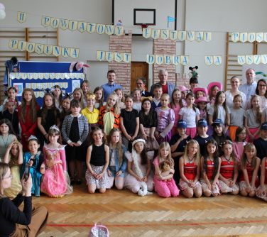 Jasna sala gimnastyczna. Grupa osób dzieci i dorośli pozują. Nad nimi na linach tytuł festiwalu