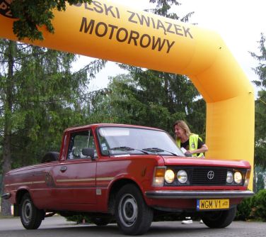 Dzień. Czerwony zabytkowy samochód stoi na linii mety. Nad nim żółtym balon. W tle drzewa