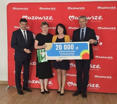 W jasnym pomieszczeniu, na drewnianej klepce dwóch mężcyzn i dwie kobiety. Trzymaja baner z kwota 20000. W ich tle czerwony...
