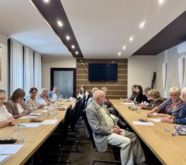Jasne pomieszczenie. Przy dwóch stołach ustawionych wzdłóż siedzą osoby