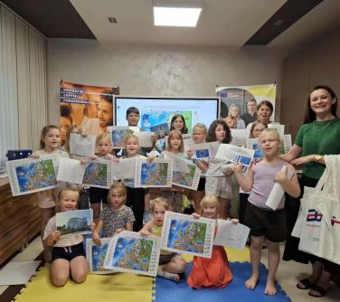 Jasne pomieszczenie. Grupa dzieci z mapami w dłoniach. Po prawej usmiechnięta kobieta w zielonym swetrze i zawieszonymi na...