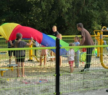 Dwór, słoneczny dzień. Na piasku na ogrodzonym placu zabaw mężcyzzna prowadzi animacje dla dzieci. W tle zielone drzewa